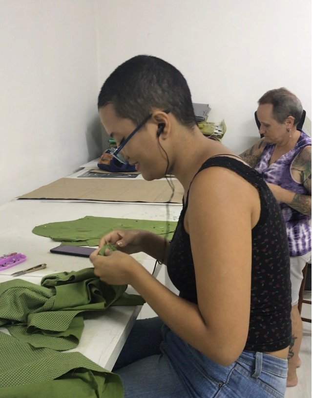 Alunas da Escola de Moda Paulistana sentadas à mesa realizando o alinhavo de peça para prova de roupa sob medida.